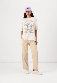 Cream oversized t-shirt with palm tree graphic, light beige corduroy trousers, and white sneakers. Purple cap with embroidered detail.