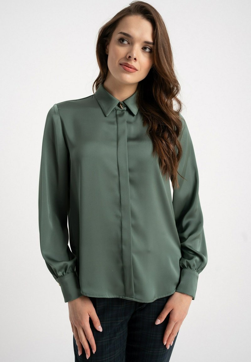 Satynowa zielona bluzka z kołnierzykiem, plisą na guziki i marszczonymi mankietami; gładka tekstura z delikatnym połyskiem.