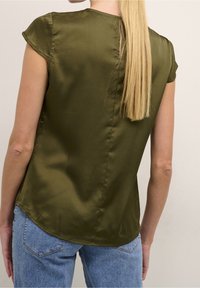 Femme aux longs cheveux blonds portant un chemisier en satin vert olive à manches courtes et un jean bleu clair, vue de dos sur un fond uni.