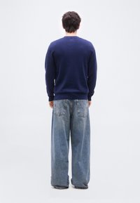 Άνδρας που στέκεται και κοιτάζει έναν λευκό τοίχο, φοράει μπλε σκούρο πουλόβερ και χαλαρά ανοιχτό μπλε τζιν με ορατές πίσω τσέπες.