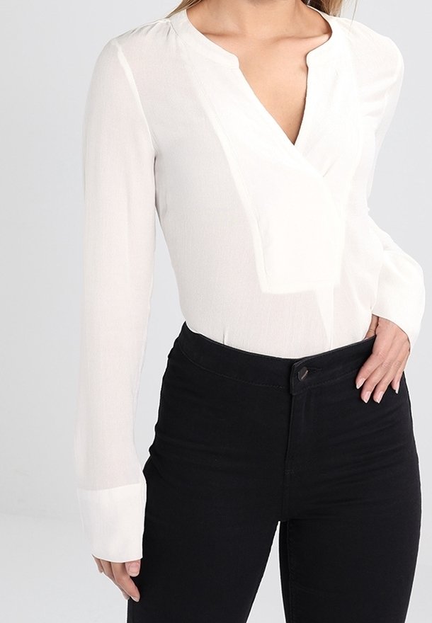 Femme portant un chemisier blanc à manches longues avec un col en V et un pantalon noir taille haute, main posée sur la hanche.