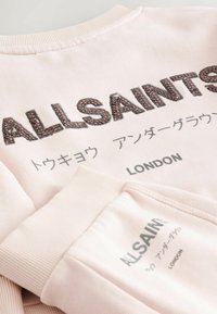 Ljusrosa sweatshirt med broderad text "ALLSAINTS" och grå tryckta detaljer. Ribbad krage och ärmavslut; mjuk tygtextur.