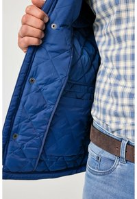 Wnętrze niebieskiej pikowanej kurtki ukazujące gładką teksturę, zatrzaski i wewnętrzną kieszeń. Noszona na koszuli w kratę i dżinsach.