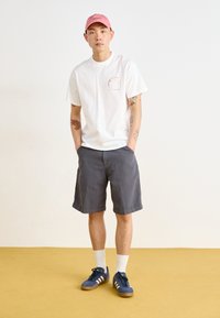 Hvit kortermet t-skjorte med liten rosa logo, grå shorts, blå joggesko med hvite striper og rosa caps. Enfarget bakgrunn.