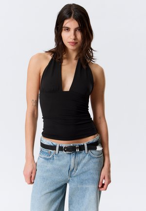 Õlgadeni tume juustega naine, kes kannab musta kaelaümbrisega topi ja helesiniseid teksapükse musta vööga, seisab ühevärvilise tausta ees.