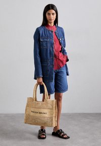 Denim jack met patches, rode shirt, denim shorts en een geweven strooien tas in natuurlijke kleur met zwarte tekst. Zwarte sandalen met bandjes.