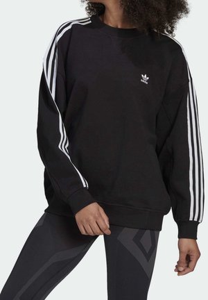 Personne portant un sweat-shirt Adidas noir avec des bandes blanches sur les manches et des leggings sombres à motifs sur un fond uni.