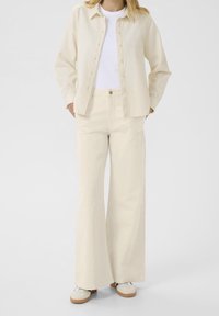 Chemise en lin de couleur crème, style boutonné ; associée à un pantalon crème taille haute, coupe à jambes larges. Des baskets blanches décontractées complètent la tenue.