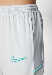 Pantaloni sportivi grigio chiaro realizzati in materiale traspirante. Presentano tasche laterali e un logo Nike ricamato turchese sul davanti.