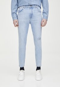 Jeans de mezclilla en azul claro con corte slim, que presentan un detalle de rodilla desgastada y un diseño clásico de cinco bolsillos, acompañados de zapatillas blancas.