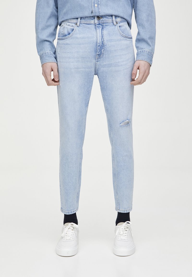 Jeans de mezclilla en azul claro con corte slim, que presentan un detalle de rodilla desgastada y un diseño clásico de cinco bolsillos, acompañados de zapatillas blancas.