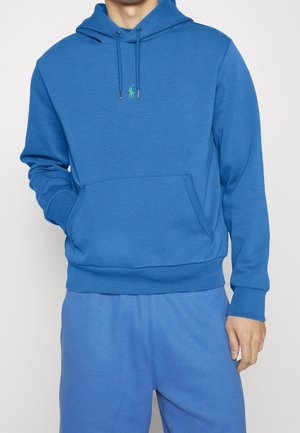Persoon draagt een blauwe hoodie met een voorkantzak en trekkoorden, gecombineerd met bijpassende blauwe broek, staand met één hand in de zak.