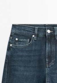 Donkere blauwe denim jeans taille gedeelte met voorste knoop, ritssluiting, lussen voor riem, en drie zichtbare zakken met klinknagels op een witte achtergrond.