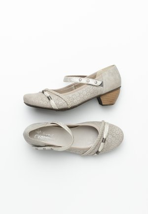 Chaussures pour femmes beiges à talons bas avec détails perforés, fermeture à bride métallique et bandes décoratives tressées sur fond blanc.