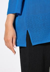 Top blu lavorato a maglia con maniche a tre quarti e uno spacco laterale, caratterizzato da un pannello in rete testurizzato all'orlo. Abbinato a pantaloni neri testurizzati.