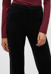 Mustad mustritepingud laieneva jala disainiga, millel on soonikkoes talje, nööbiklipp ja tekstuuriline pind. Näha on punane pikakäistega pluus.