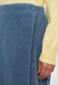 Modrá kordurová sukňa s gombíkom potiahnutým látkou v páse, nosená s žltým svetrom s dlhým rukávom, ruka odpočíva po boku.