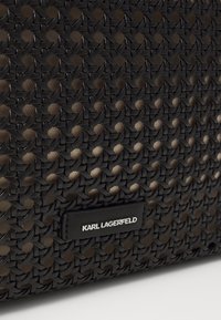 Austas juodas tekstilinis krepšys, pasiūtas su lygaus odos žyme, ant kurios sidabro spalva užrašyta "KARL LAGERFELD". Dizainas yra geometrinis su tekstūrizuota paviršiumi.