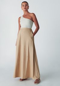 Maxi gonna beige chiaro con tasche laterali, abbinata a un top bianco a una spalla con costine. Il tessuto sembra morbido e leggero.