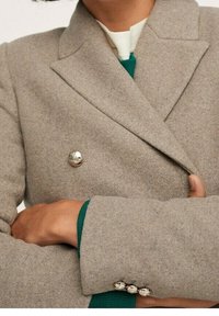 Blazer doppiopetto in lana grigia chiaro con bottoni metallici. Caratterizzato da rever a increspatura e tessuto strutturato. Maglione verde visibile sotto.