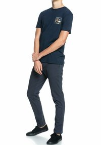Mörkblå T-shirt med ficklogotyp, matchad med mörkgrå byxor. Modellen har på sig svarta sneakers. Enkelt design och bekväm passform.