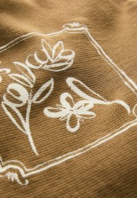 Textile marron ornée de broderies florales blanches entourée d'une bordure rectangulaire ; tissu texturé avec un motif de tissage visible.