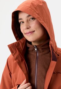 Giacca impermeabile rosso-marrone con cappuccio, dotata di chiusura con zip, dettagli in Velcro e uno strato interno strutturato. Logo visibile sul lato.