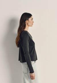 Ženska z dolgimi rjavimi lasmi stoji v profilu, oblečena v temno dolg rokav in svetlo modre kavbojke, ob preprosti bež steni.