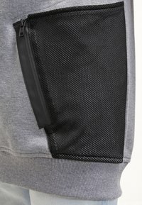 Šedá mikina s texturovanou černou síťovanou kapsou se zipem. Kapsa je velká a obdélníková, což kontrastuje s hladkým materiálem.