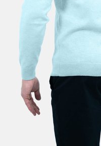 Lichtblauwe gebreide trui met geribbelde manchetten, van dichtbij naast een hand getoond, gecombineerd met zwarte broek. Zachte textuur, strak ontwerp.