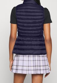 Ärmellose marineblaue Steppweste mit horizontalen Nähten, hohem Kragen und glatter Textur, kombiniert mit einem hellen, karottengerüschten Rock.