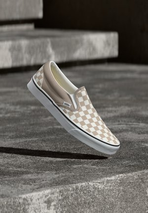 Beige and white checkerboard slip-on Vans shoe hovering above textured concrete stairs outdoors.