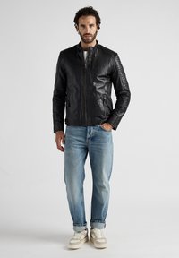 Zwarte leren bomberjack met gequiltte schouderdetails, vooraan met ritssluiting en zijzakken, gedragen over lichtblauwe jeans en witte sneakers.