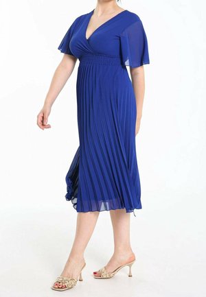 Femme portant une robe plissée bleu roi mi-longue avec des manches papillon transparentes et des sandales à talons beiges, debout devant un fond blanc.