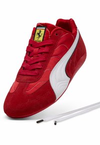 Rote Wildleder- und Stoff-Sneaker mit Ferrari-Logo, weißer Puma-Streifen und losen weißen Schnürsenkeln auf einem weißen Hintergrund.