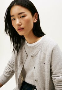 Lichtgrijze gebreide cardigan met een ronde halslijn en knopen, met ribbels aan de mouwen en een klein logo op de linkerkant van de borst.