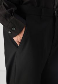 Schwarze Hosen aus glattem Stoff, ausgestattet mit einer Seitentasche und einem Gürtelschlaufe. Der sichtbare Hemdsärmel zeigt markante Knöpfe.