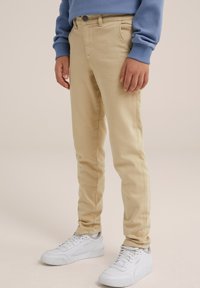 Bež pamut twill nadrág, karcsú fazonnal, elülső zsebekkel és gombos záródással. Fehér tornacipővel és kék pulóverrel párosítva.