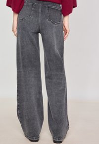 Jean en denim noir à jambes larges avec une finition texturée, comportant deux poches arrière, une taille haute et des ourlets droits. Amélioré par des détails de couture subtils.