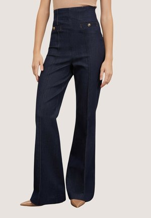 Pantalon bleu foncé taille haute à jambes larges avec détails de couture devant et pattes de taille boutonnées, porté avec des chaussures pointues beige.