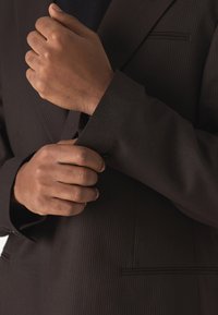 Ruskea raidoitettu bleiseri, jossa on muotoiltu istuvuus; kädet kiinnittävät hihansuut, paljastaen tekstuurin ja hienovaraisen taskuyksityiskohdan.