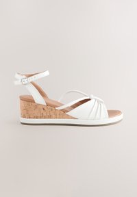 Sandale à talon compensé blanc avec talon en liège, détail de sangle nouée et bride de cheville réglable. Texture lisse et semelle intérieure rembourrée.