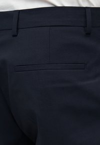 Σκούρο μπλε παντελόνι με ζώνη και μονή τσέπη πίσω, φορεμένο με λευκό πουκάμισο μέσα από το παντελόνι.