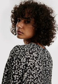 Schwarze Bluse mit floralen Mustern in Weiß, strukturiertem Stoff, Rundhalsausschnitt und einer Knopfdetails hinten. Lockige Haare sind sichtbar.