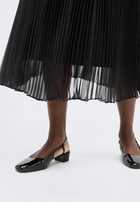 Κάτω άκρα με λαμπερά μαύρα πέδιλα slingback και διάφανο μαύρο πλισέ midi φούστα σε λευκό φόντο.