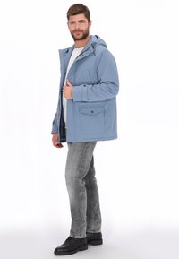 Chaqueta con capucha de color azul claro con cremallera frontal, dos bolsillos en el pecho y tela texturizada. Se lleva sobre una camiseta gris y combinada con vaqueros grises.