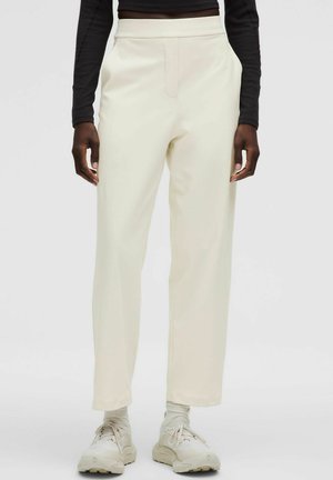 Osoba ubrana w kremowe spodnie o prostych nogawkach, białe trampki i czarną bluzkę z długim rękawem, stojąca na jednolitym tle.