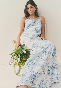 Rochie maxi florală dintr-o țesătură moale, cu un fundal alb și trandafiri ghinți, bretele subțiri și un design fluid. Mână ținând un buchet.