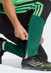 Zelene športne hlače z teksturiranim vzorcem, črnimi poudarki in zadrgami na gležnjih. Nošene s črnimi nogometnimi škornji z izrazito zelenimi poudarki.