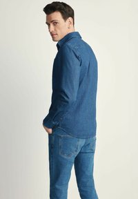 Koszula dżinsowa w ciemnoniebieskim kolorze, z długimi rękawami, zapinanymi mankietami i kołnierzykiem. Połączona z dopasowanymi ciemnoniebieskimi dżinsami z tylnymi kieszeniami.